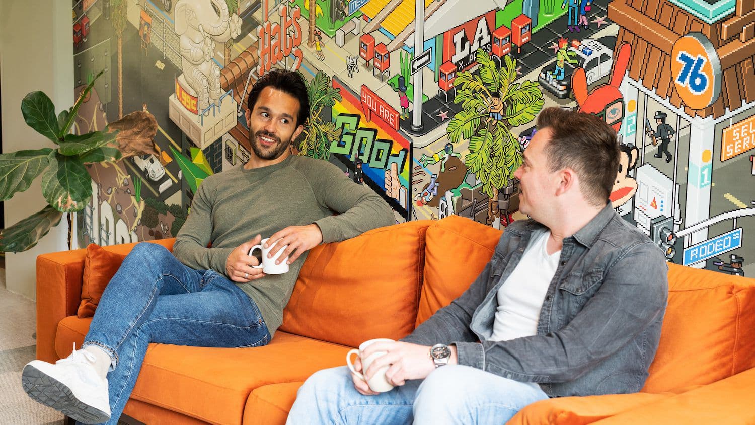 Twee collega's in gesprek op een oranje bank voor een muur met een kleurrijke illustratie
