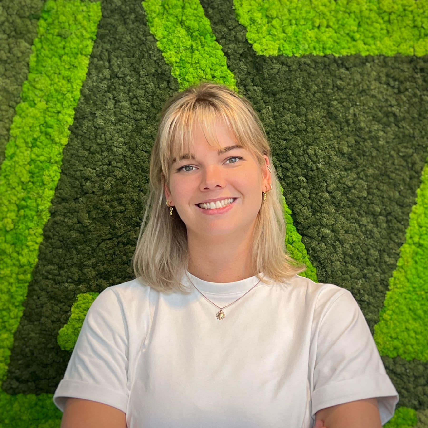 Portret van Rosina Meijnen, lachend met gekruiste armen voor een groene mosmuur
