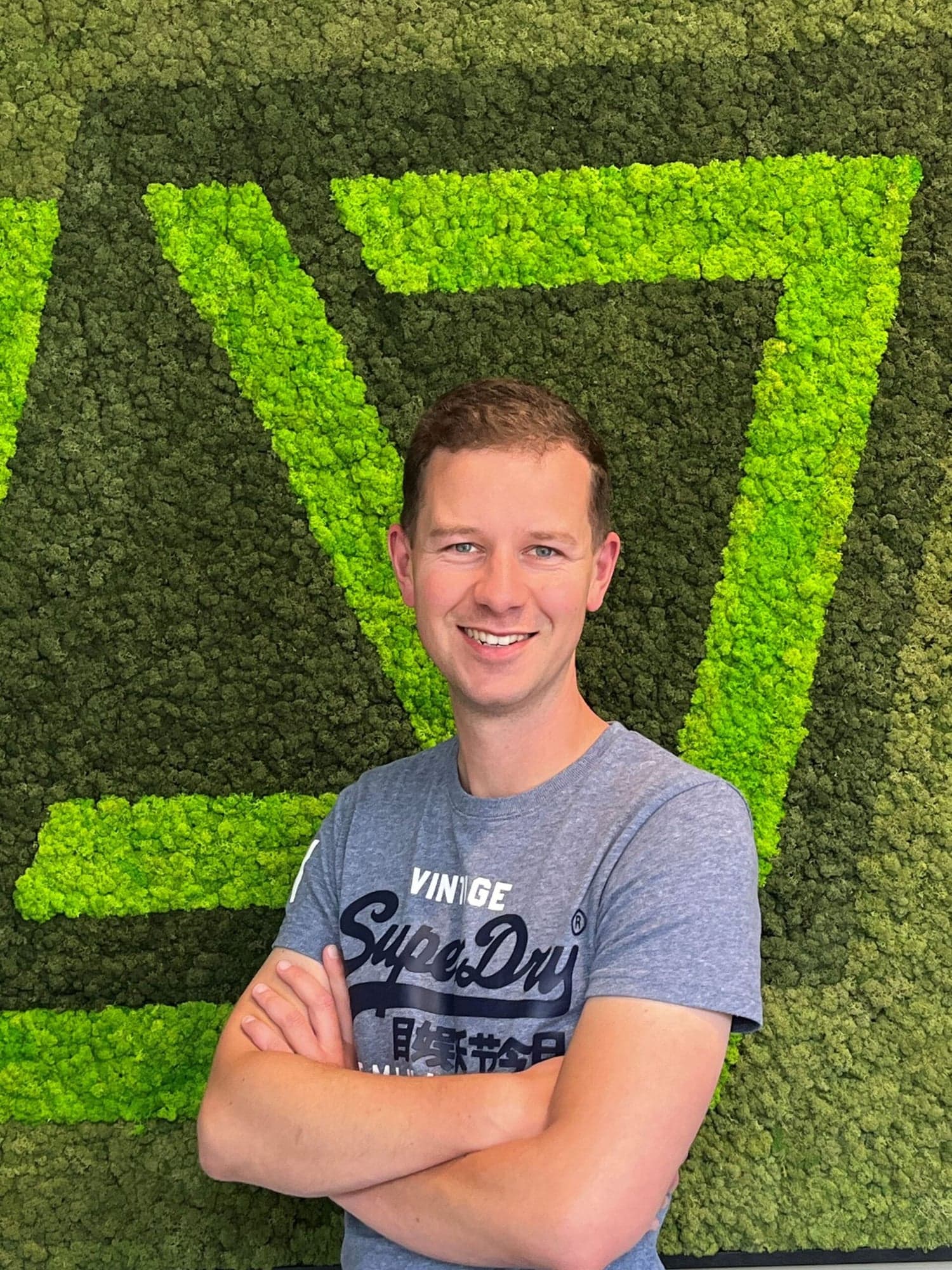 Portret van Michael Klaassen, lachend met gekruiste armen voor een groene mosmuur