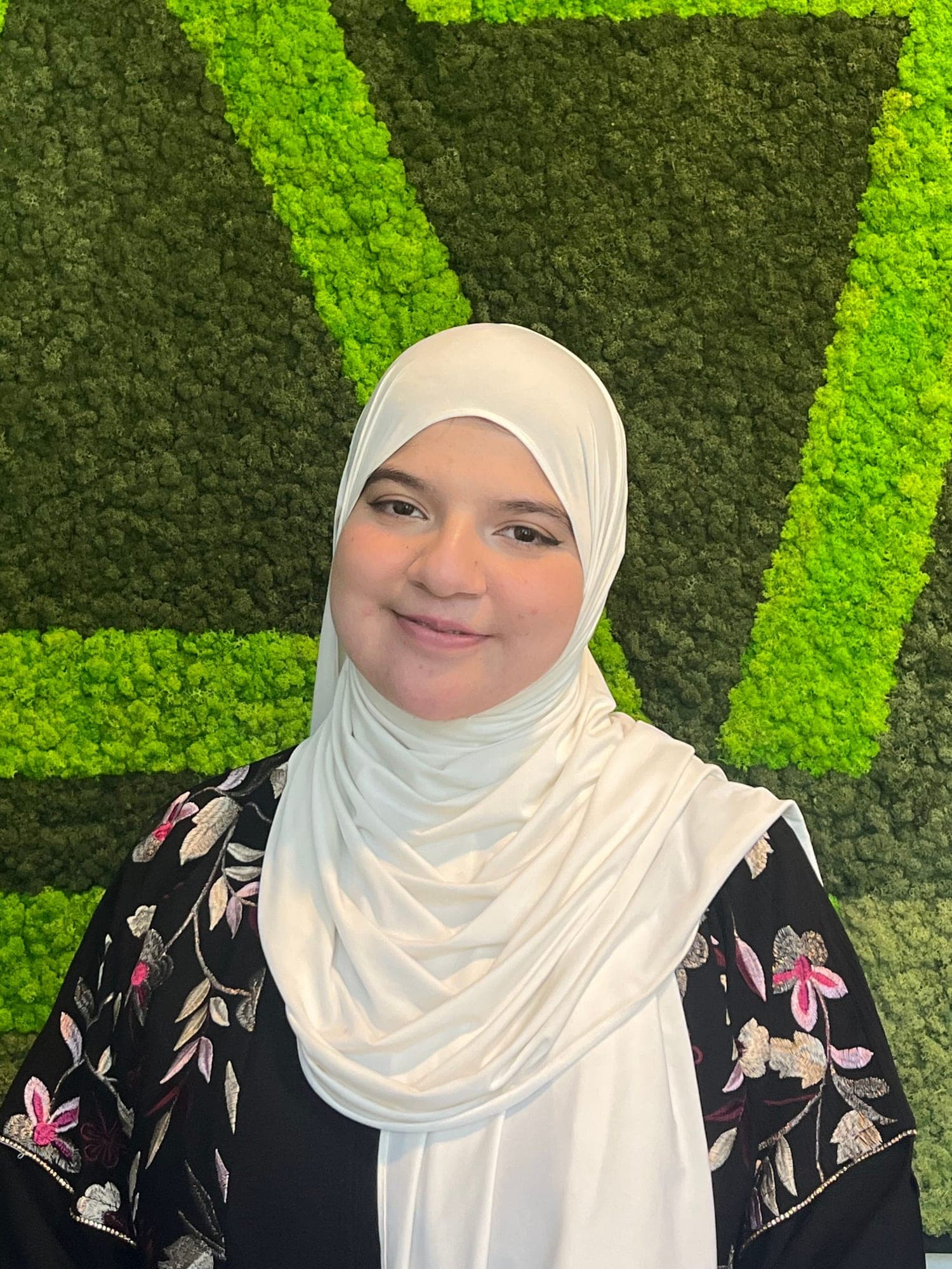 Portret van Hijab Muhammad, glimlachend voor de groene mosmuur met het JoinSeven-logo