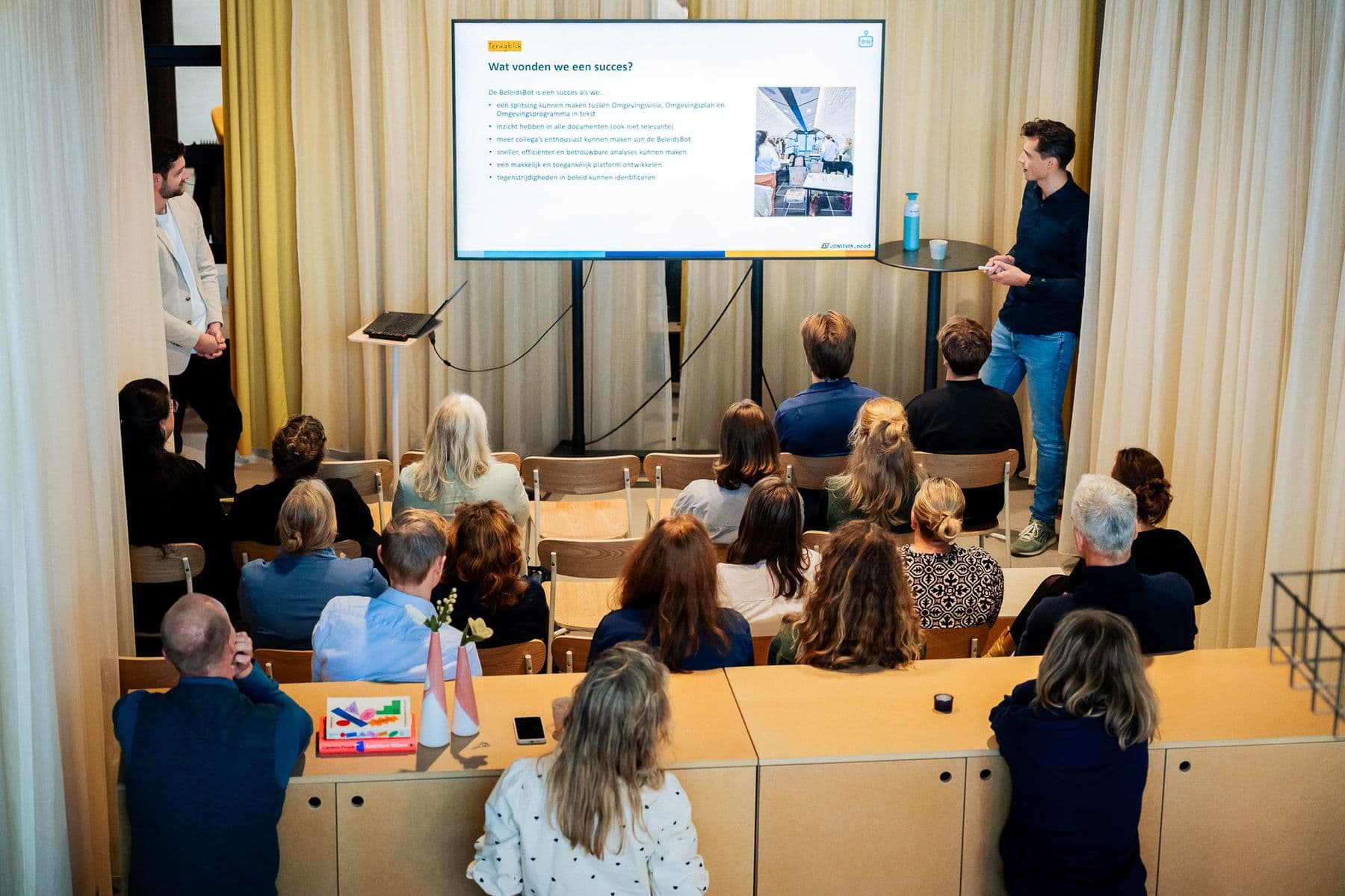 Twee collega's van JoinSeven presenteren op een groot scherm de resultaten van een project aan een zaal vol vakgenoten